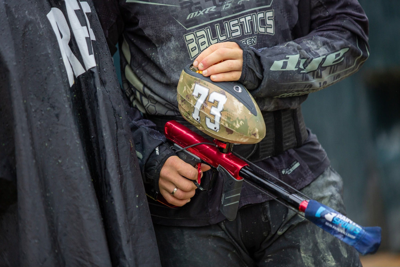 Paintball-Spieler mit rotem Paintball-Markierer in Deckung, hält den Markierer bereit hinter einer schwarzen Spielfeld-Barriere.