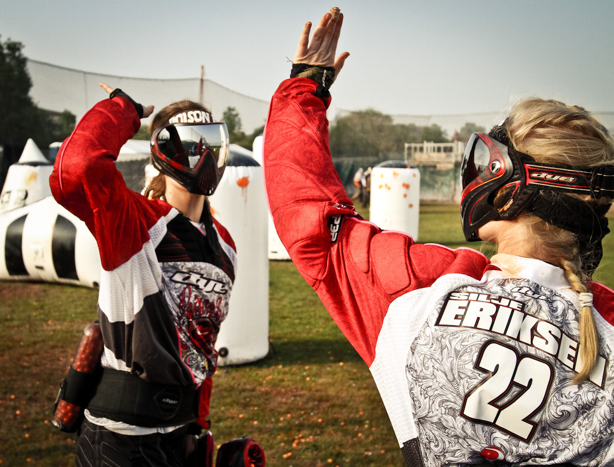 Paintball-Spieler mit Markierern und Paintball-Masken feiern einen Treffer auf dem Spielfeld mit erhobenen Händen.